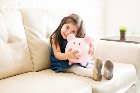 Portrait of beautiful little girl holding a big piggy bank in hands while sitting on sofa in living roomの写真素材