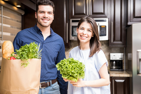 Beautiful latin couple standing together in their kitchen with grocery shopping bag and fresh vegetablesの写真素材