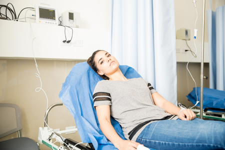Young latin woman lying on hospital bed with an IV drip looking worried about her health and recoveryの写真素材
