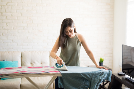 Cheerful young housewife wearing casuals ironing men's shirt on ironing boardの写真素材