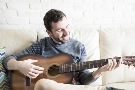 Portrait of handsome young latin man playing a guitar and smiling at homeの写真素材