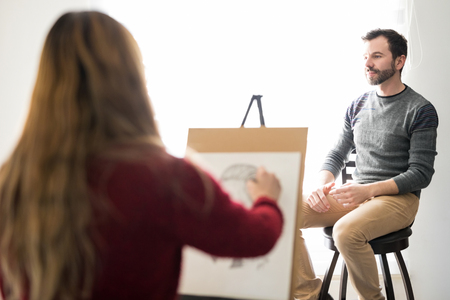 Rear view of a young female artist sketching a man sitting on stool indoorsの写真素材