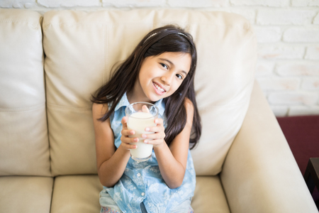 Happy child girl drinking milk in glass while sitting on couch at homeの写真素材