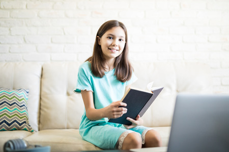 Smiling school girl completing homework by sitting on couch in living room with laptop and...