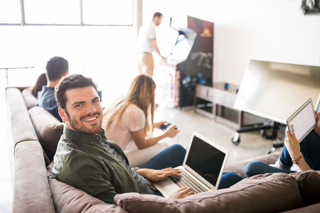 Portrait of good looking latin man using laptop in lounge room in office with colleagues relaxing...