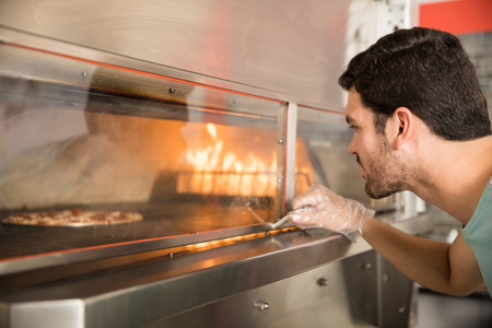 Portrait of handsome chef wearing gloves holding oven handle checking on pizzaの写真素材