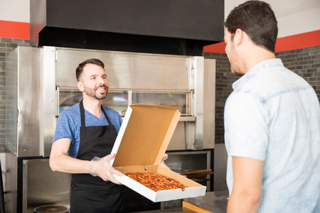Handsome young chef with bears wearing black apron uniform and disposable gloves holding open box of pizza while customer standing at counter in pizza shopの写真素材