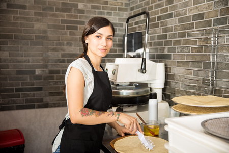 Pretty chef working in kitchen preparing raw pizza bread wearing black apron uniform in pizzeria kitchen while looking at cameraの写真素材