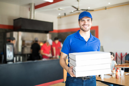 Handsome pizza delivery man courier in blue uniform with cap holding pizza boxes smiling and looking at cameraの写真素材