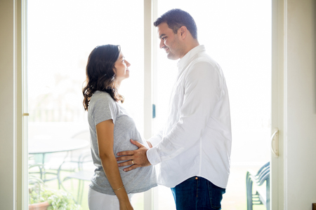 Mid adult man touching pregnant wife's belly while standing at homeの写真素材