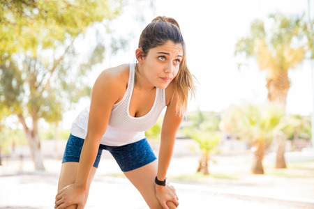 Beautiful young woman standing with hands on knees and having rest after morning run at the parkの写真素材