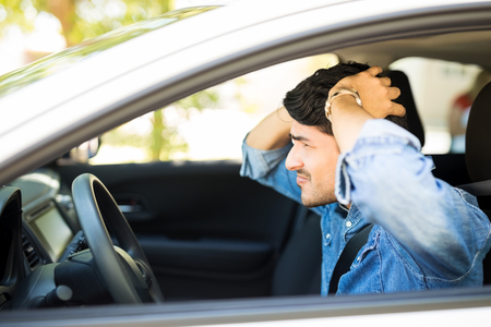 Profile view of young latin man sitting inside his car with hands on head during traffic jamの写真素材