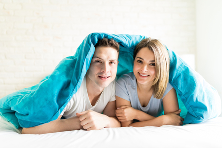Portrait of confident couple smiling while lying under blanket on bed at homeの写真素材