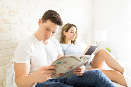 Handsome male reading newspaper by woman sitting on bed at homeの写真素材