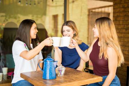Pretty hispanic female friends having fun and making a toast with coffee cups at restaurantの写真素材