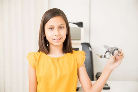 Portrait of cute girl standing in yellow shirt holding eye test glasses in optical store while looking at cameraの写真素材