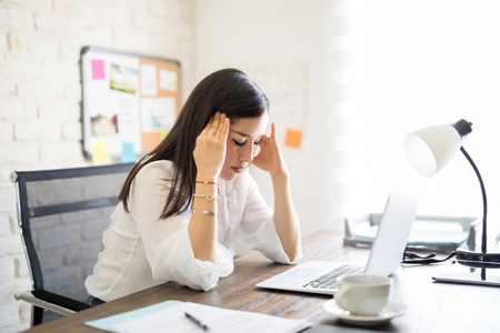 Woman in her 30s sitting at the work desk holding her head, businesswoman suffering from headache.の写真素材