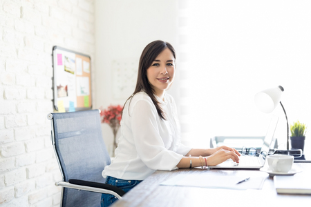 Portrait of attractive young hispanic woman sitting at her desk table working on laptop and making an eye contactの写真素材