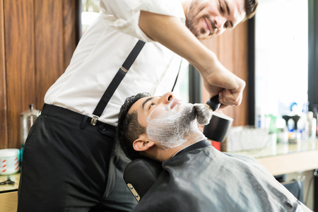 Male barber rubbing shaving foam on client's face with brush in shopの写真素材