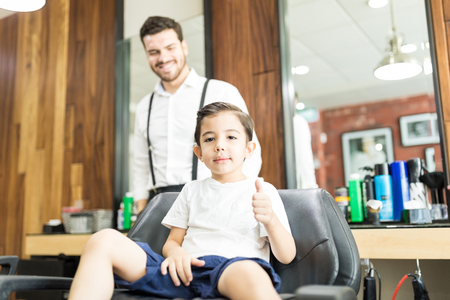 Cool boy showing thumbs up while sitting on chair at salonの写真素材