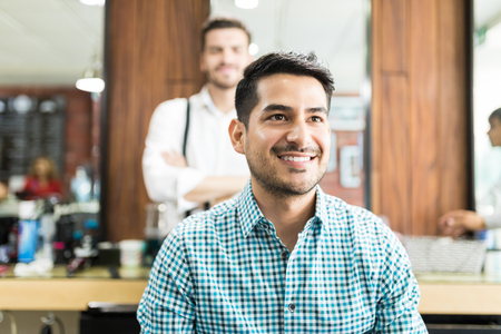 Smiling young man looking away after haircut with barber in background at salonの写真素材