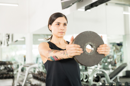 Confident young woman exercising with barbell plate during gym classの写真素材