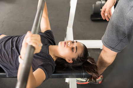Young sporty woman doing the bench press by instructor in gymの写真素材