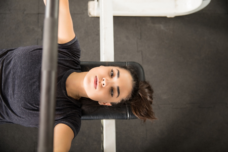 Directly above shot of determined woman pumping muscles with barbell on bench press in gym