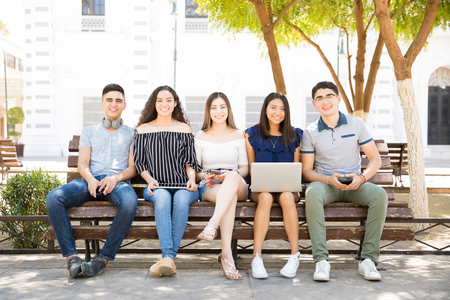 Portrait smiling five teens sitting on outdoor bench with laptop, digital tablet and mobile phonesの写真素材