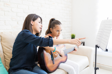 Mid adult teacher pointing at musical notes while teaching girl to play guitar at homeの写真素材