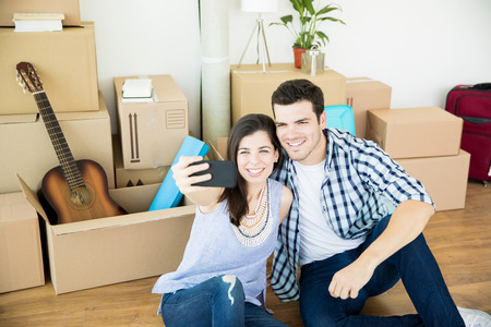 Happy mid adult man and woman taking selfie on smartphone while sitting against moving boxes at new...