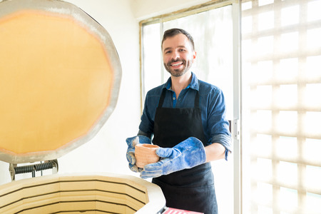Portrait of smiling mid adult owner holding freshly made cup while standing by oven in pottery...