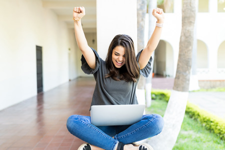 Female with arms raised screaming while using laptop on railing at campusの写真素材