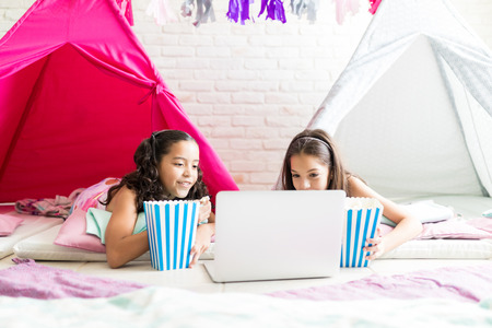 Multiethnic girls with popcorn buckets watching movie on laptop computer in tipi tents during pajama party at homeの写真素材