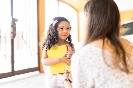 Cute little girl holding books while looking at young mother preparing for kindergartenの写真素材