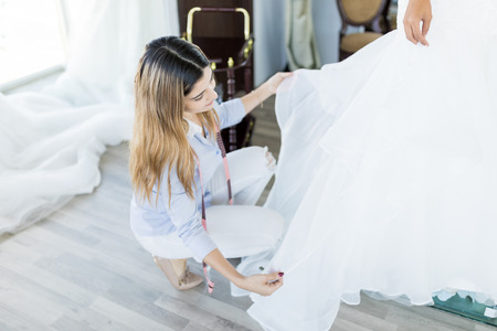 Skillful tailor examining frills of white wedding dress while crouching in shopの写真素材