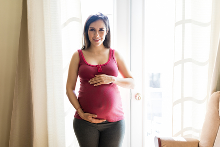 Portrait of pretty woman protecting pregnant belly while smiling at homeの写真素材