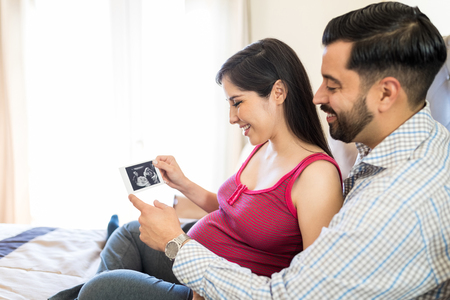 Cute mid adult couple holding ultrasound scan of unborn baby while sitting in bedroom