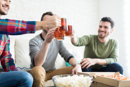 Male buddies raising beer mugs while enjoying weekend party in living roomの写真素材