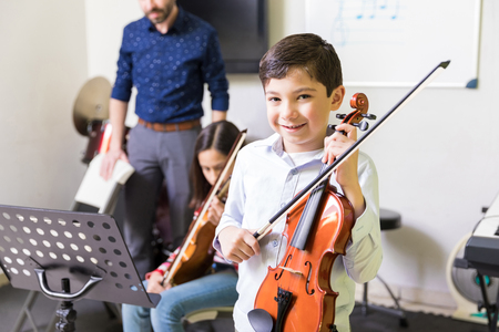 Portrait of smiling boy holding violin and bow while learning music in classの写真素材