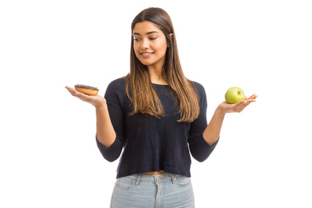 Thoughtful woman deciding between healthy apple and unhealthy donut while standing against white backgroundの写真素材