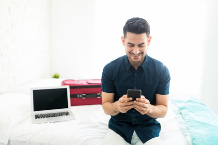 Good looking man smiling while using smartphone by laptop and luggage in bedroom