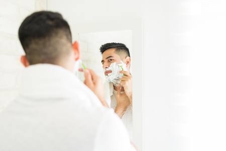 Handsome man shaving face with razor in home bathroomの写真素材