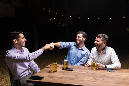 Cheerful male besties bumping their fists while enjoying beer at backyard party during night