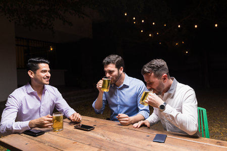 Male friends drinking beer while sitting at dining table in backyard party during nightの写真素材