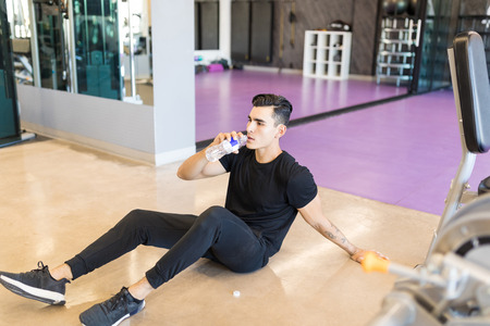 High angle view of tired young man drinking water while sitting on floor in health club