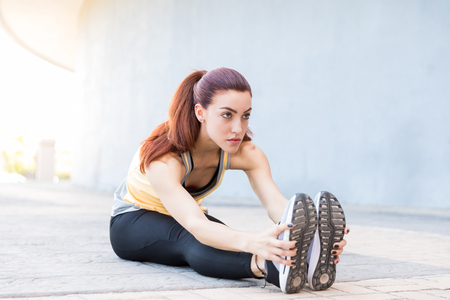 Dedicated female runner warming up while sitting on sidewalk and looking away in cityの写真素材