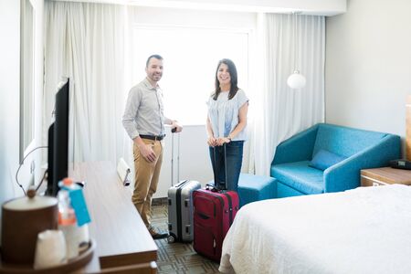 Attractive man and woman on vacation standing with suitcases by furniture while making eye contact...