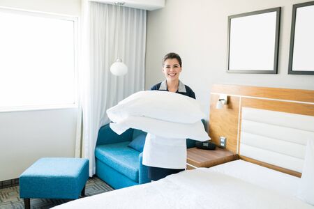 Female housekeeper carrying stack of clean pillows by bed in hotel roomの写真素材