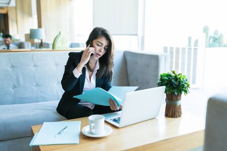 Beautiful mid adult saleswoman reading document while talking on cellphone with client in hotel lobbyの写真素材